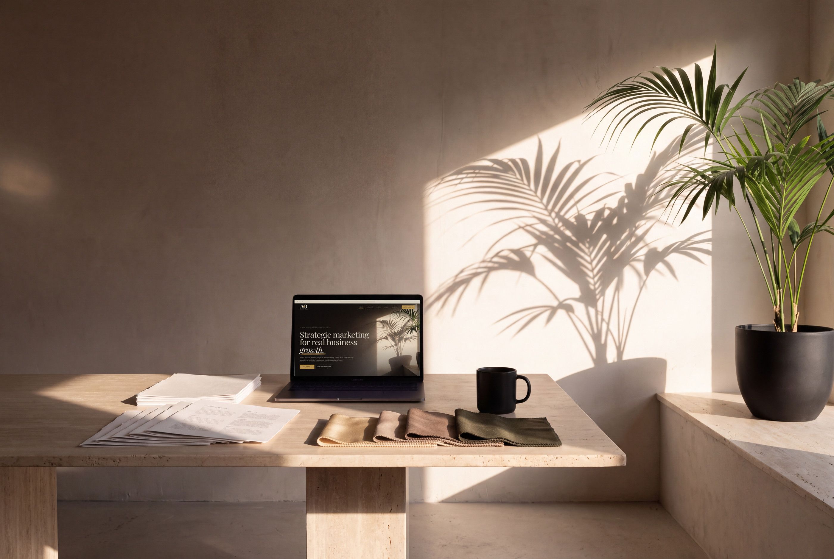 A calm studio in late afternoon sun: a travertine work table with a laptop, printed proofs, fabric swatches and a black mug, beside a palm in a black pot.